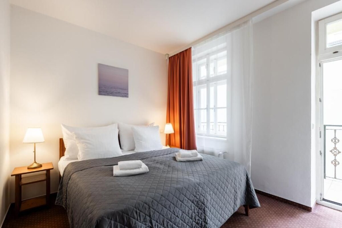 Ein Hotelzimmer mit einem Bett, zwei Nachttischlampen, einem fensterbelaubten Fenster und einer zur Außenseite führenden Tür.