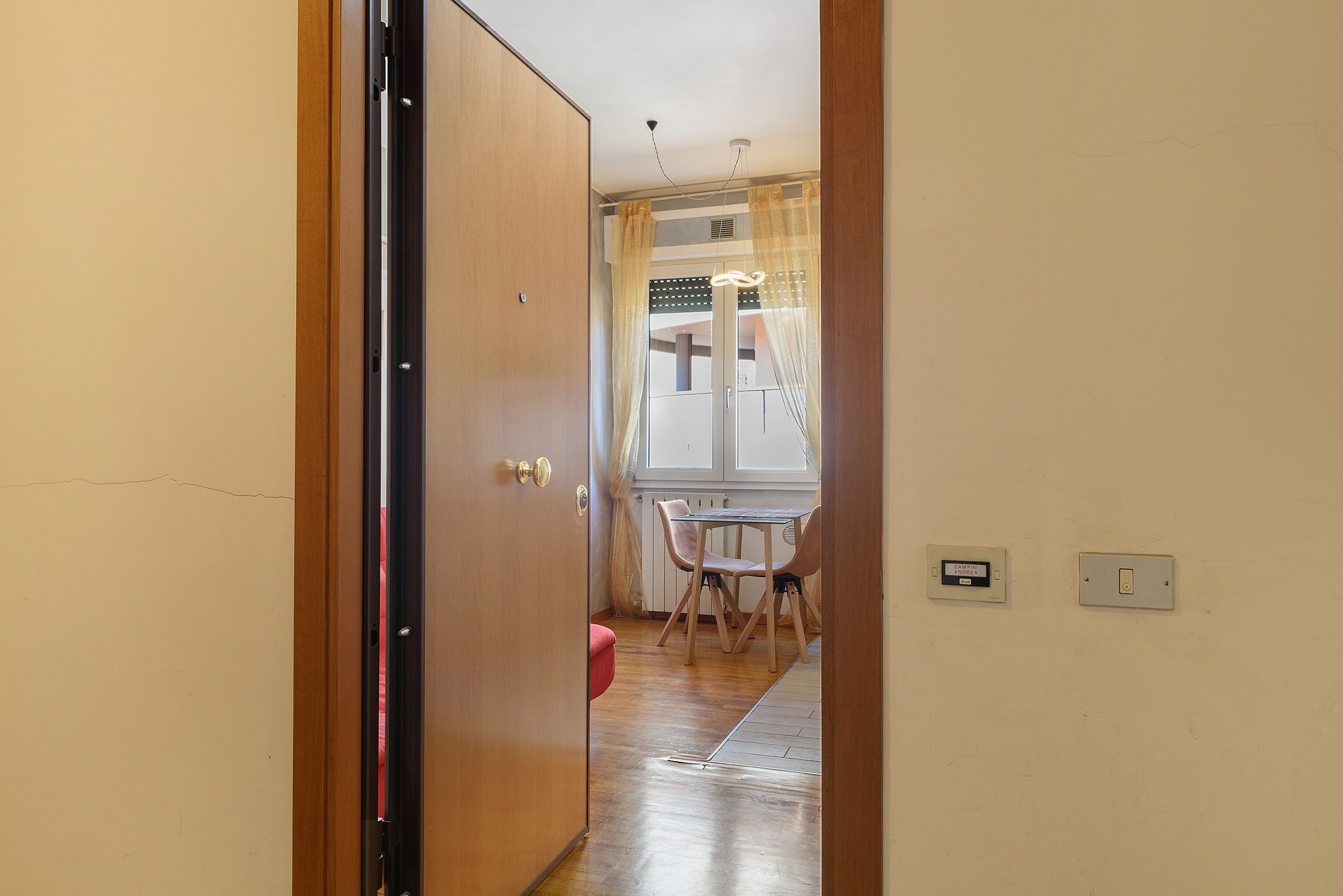 A hallway with a wooden door, a window with curtains, a table with chairs, and a light switch on the wall.
