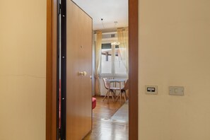 Un pasillo con puerta de madera, una ventana con cortinas, una mesa con sillas e interruptor de luz en la pared.
