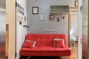 Un sofá rojo con almohadones en una habitación con pared blanca, diplomas enmarcados y un ventanal pequeño.