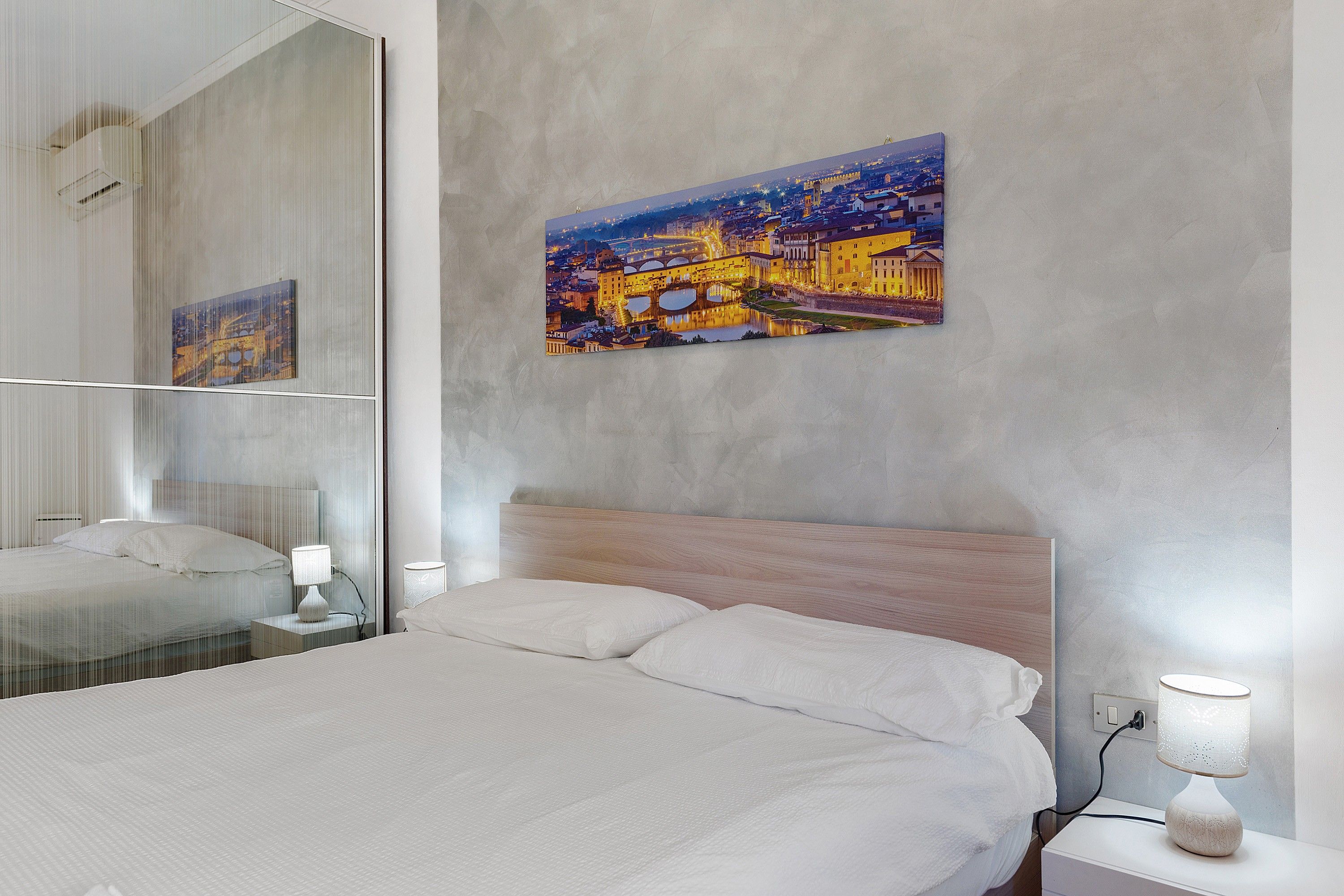 A hotel room with a large bed, a bedside table, a lamp, a mirror, and a cityscape painting on the wall.