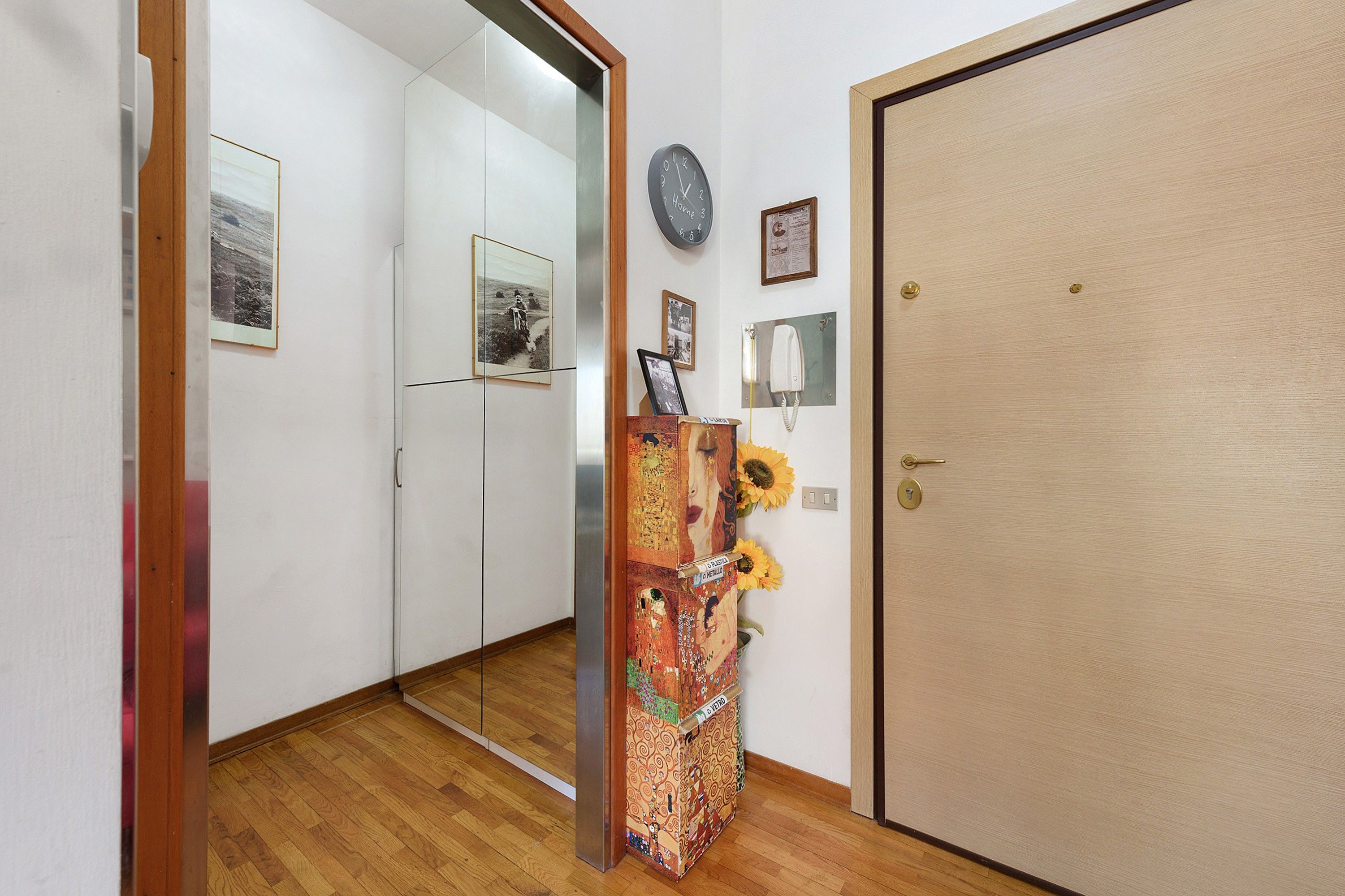 A hallway with a wooden floor, a door with a keyhole, and a wall with a clock and framed pictures.