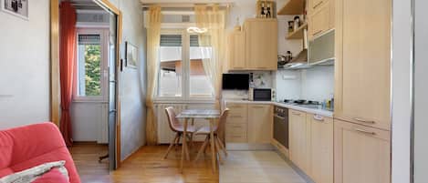 A compact kitchen with wooden cabinets, a dining table with chairs, and a red sofa in the living area.