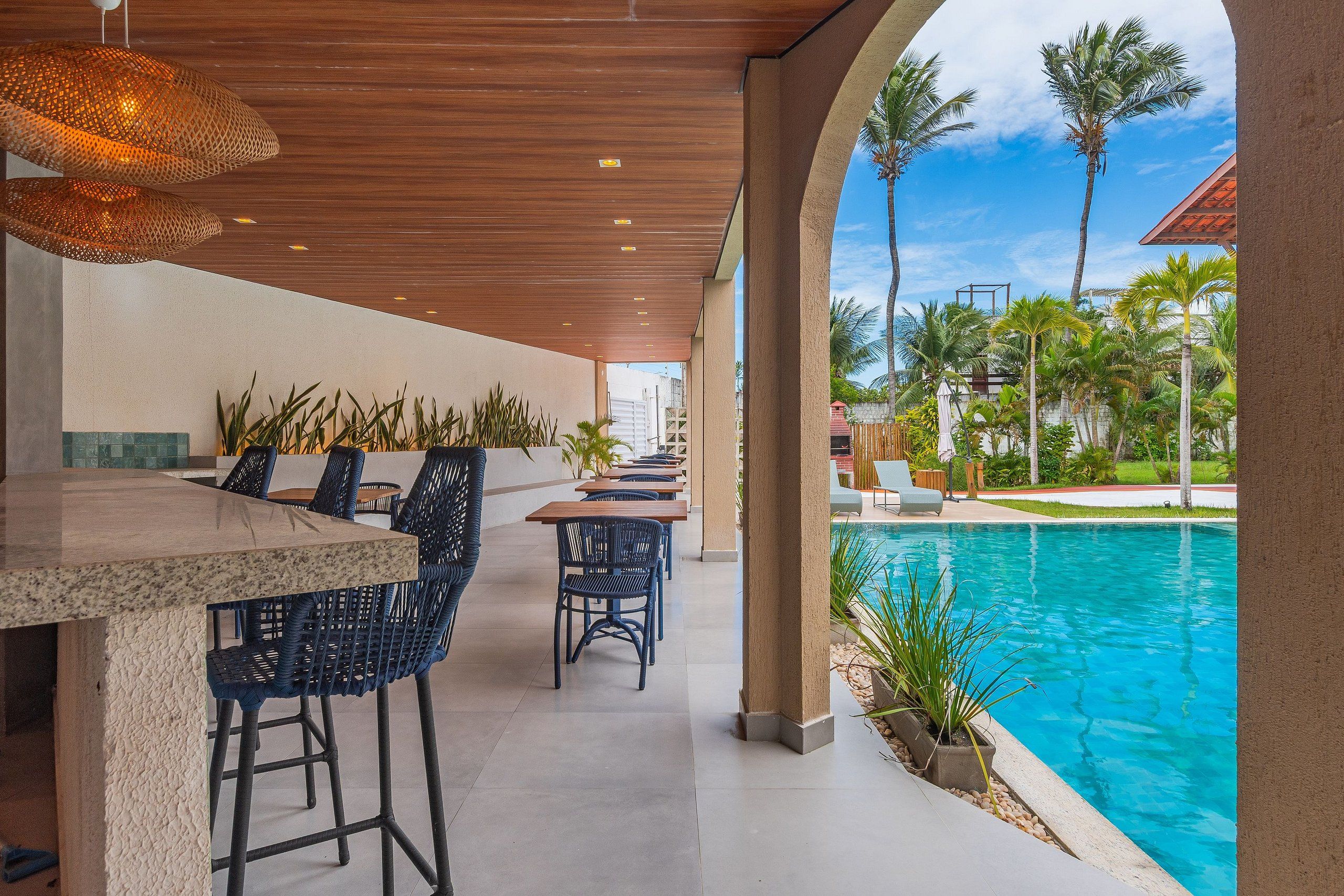An outdoor bar area with a pool view, featuring a wooden ceiling, bar stools, and lounge chairs.