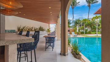 An outdoor bar area with a pool view, featuring a wooden ceiling, bar stools, and lounge chairs.