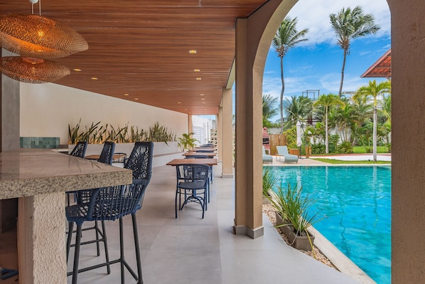 Área de bar ao ar livre com vista para a piscina, apresentando teto de madeira, banquetas e cadeiras de lounge.
