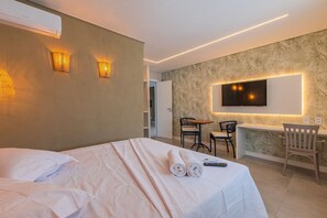 Quarto de hotel com cama, escrivaninha, cadeira, televisão e luminária na parede.