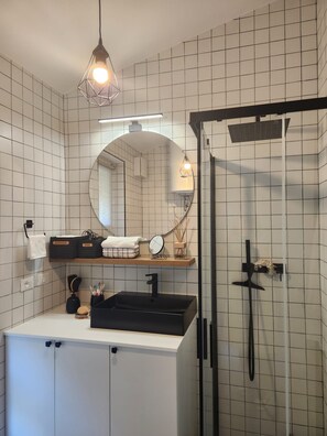 A modern bathroom with a white tiled wall, a black sink, a mirror, and a shower. - Vrbo Property (Lombardia)