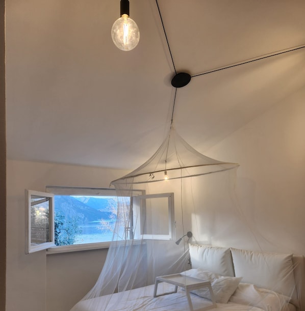 A bedroom with a bed, a bedside table, a window with a view of mountains, and a hanging light fixture. - Vrbo Property (Lombardia)
