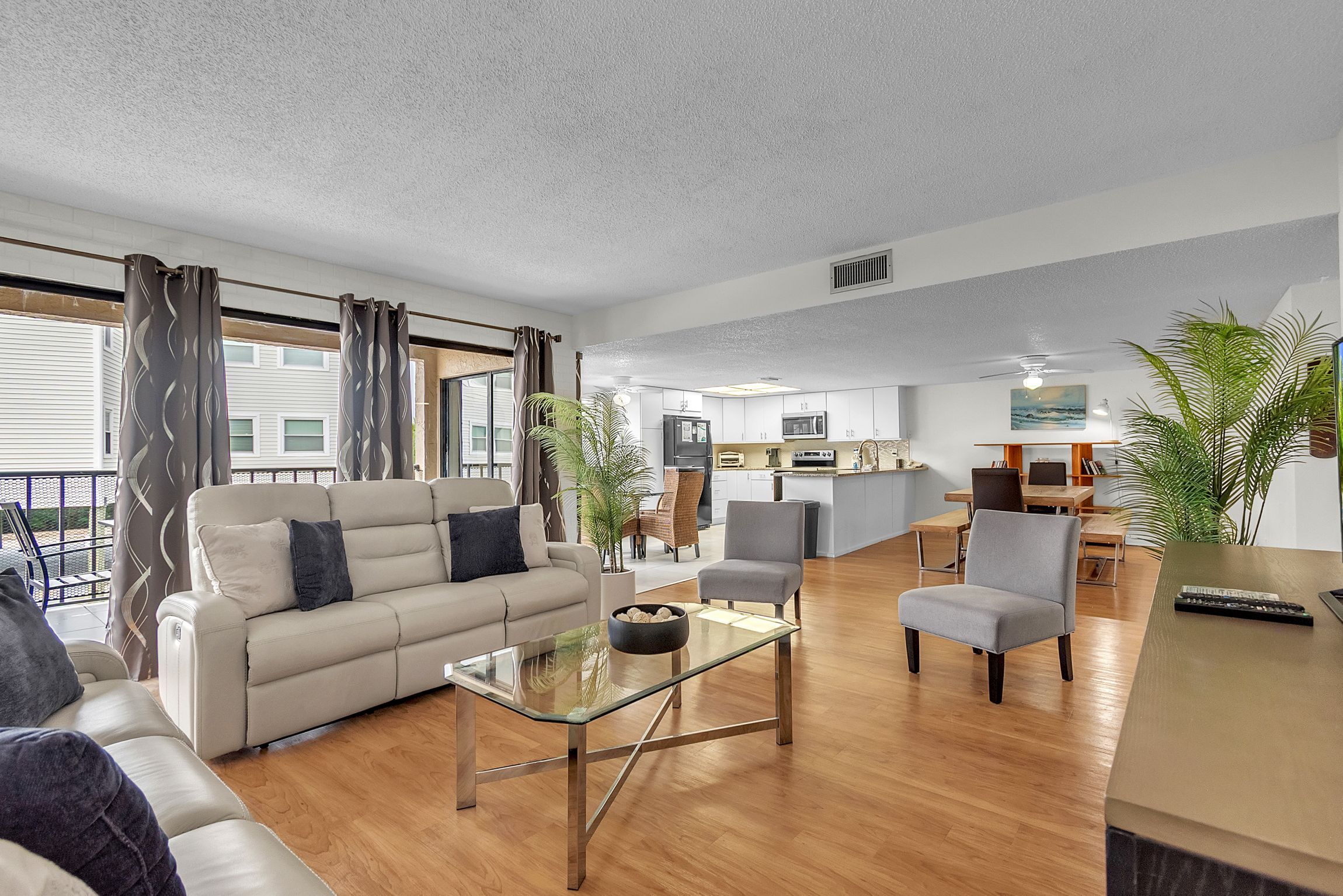 A modern living room with a sofa, armchairs, and a glass coffee table. There is a kitchen area in the background with a dining table and chairs.
