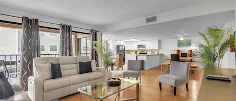A modern living room with a sofa, armchairs, and a glass coffee table. There is a kitchen area in the background with a dining table and chairs.