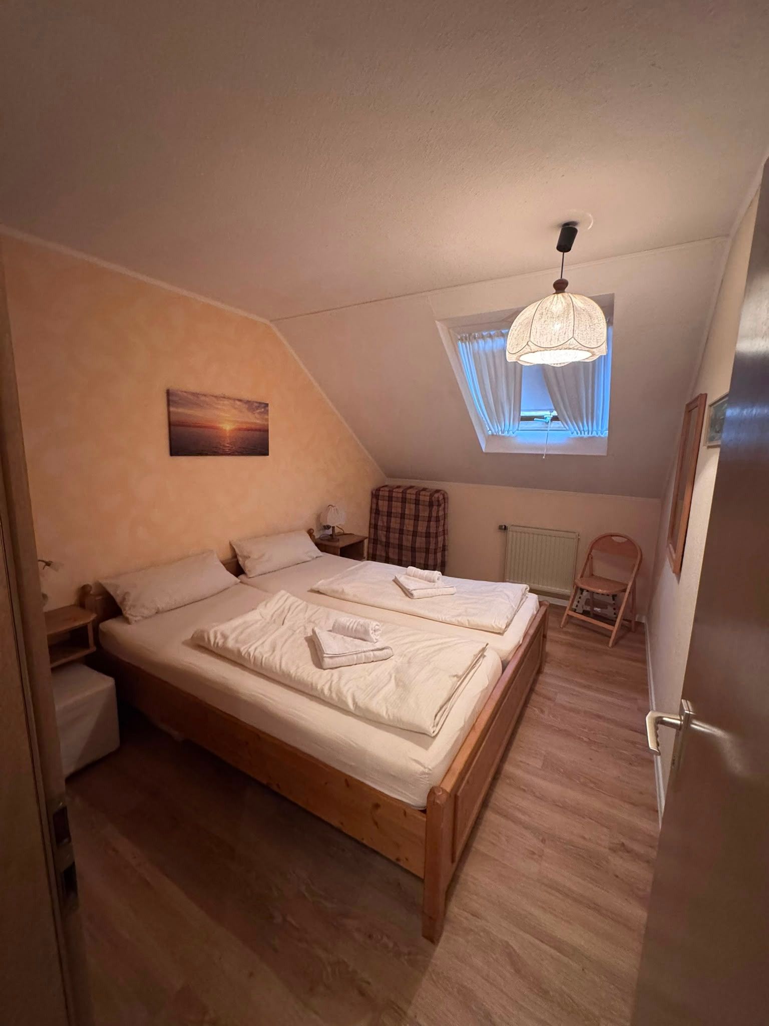 A bedroom with a wooden bed, a nightstand, a chair, a window with curtains, and a painting on the wall.