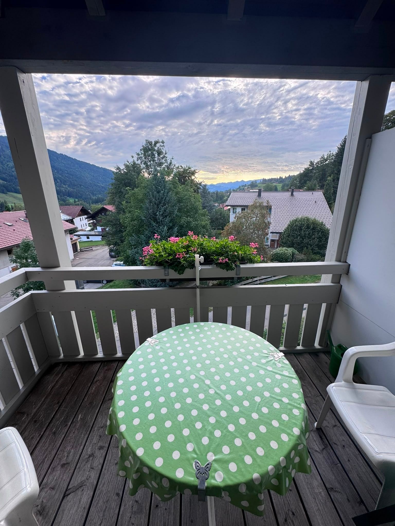 Ein Balkon mit einem runden Tisch, der mit einer grün-weiß gepunkteten Tischdecke bedeckt ist, weißen Stühlen und Blick auf eine gebirgige Landschaft.