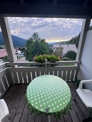 Un balcón con una mesa redonda cubierta con un mantel verde de lunares, sillas blancas y vistas a un paisaje montañoso.