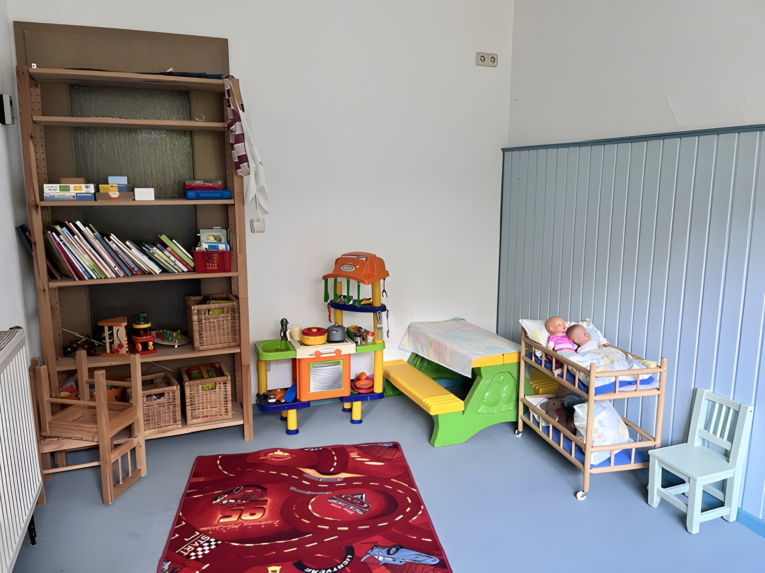 Ein Kinderzimmer mit Bücherregal, Spielküche und einem Bett mit einer Puppe.