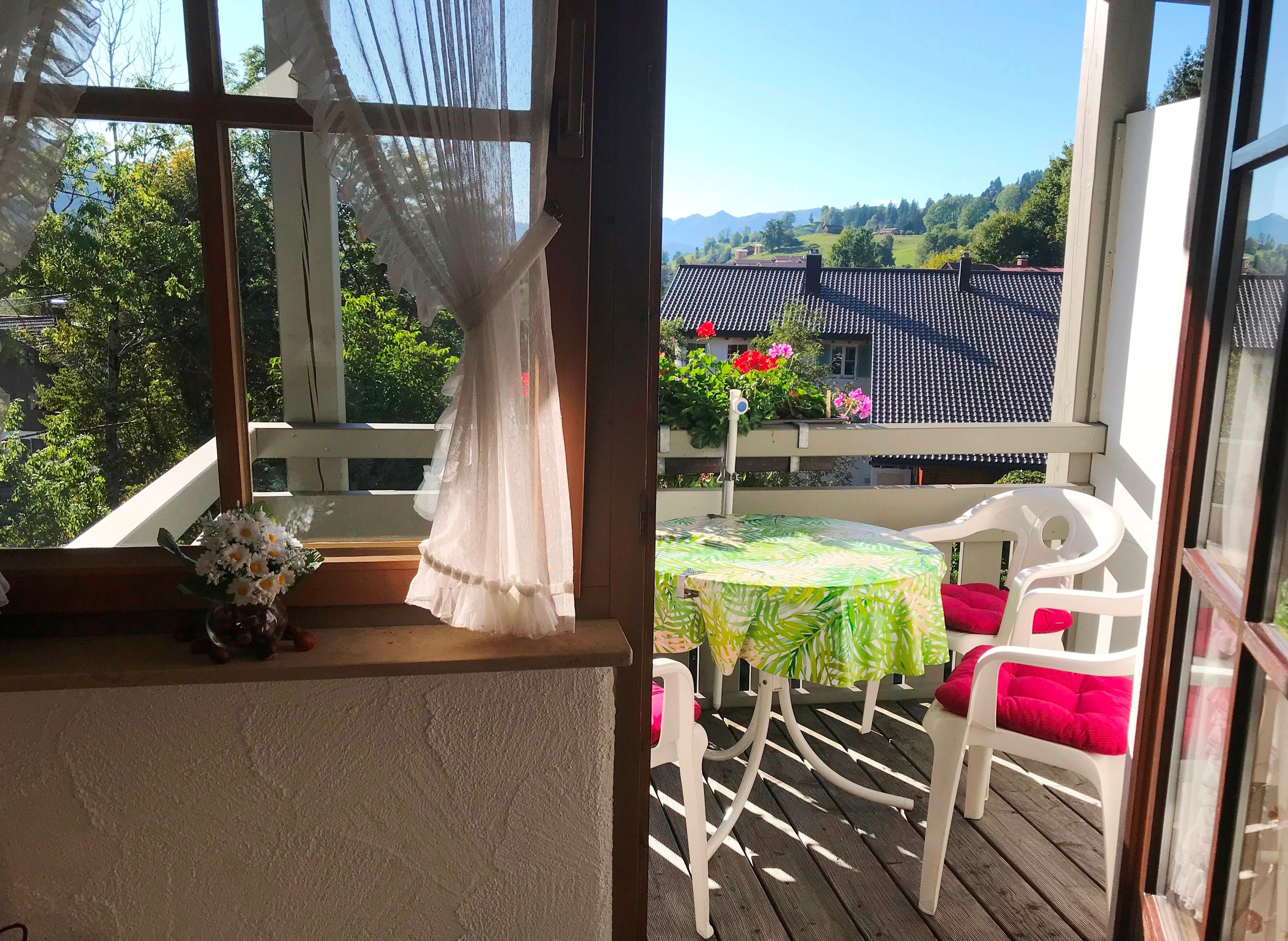 Ein Balkon mit Tisch und Stühlen, Blick auf ein Haus mit Ziegeldach und grüne Bepflanzung im Hintergrund.
