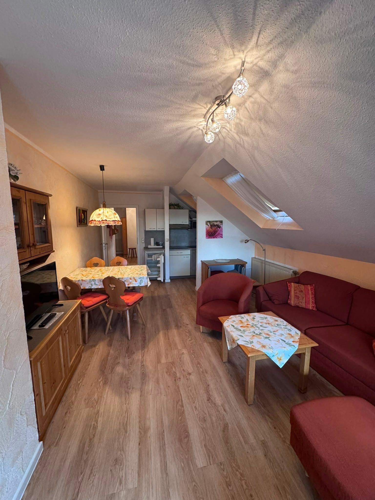 A compact living space with a dining area, a red sofa, a small table, and a kitchen in the background.