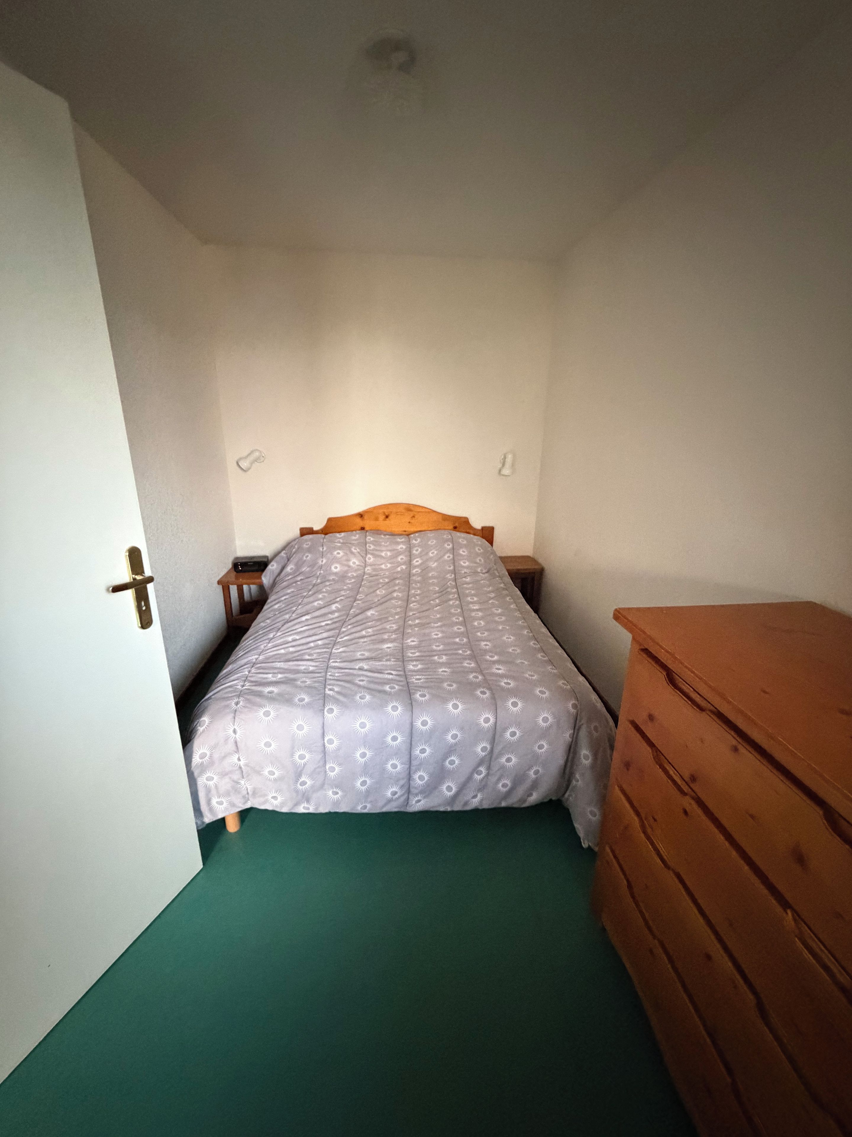 Una habitación pequeña con cama individual, cabecera de madera, alfombra verde y cómoda de madera.