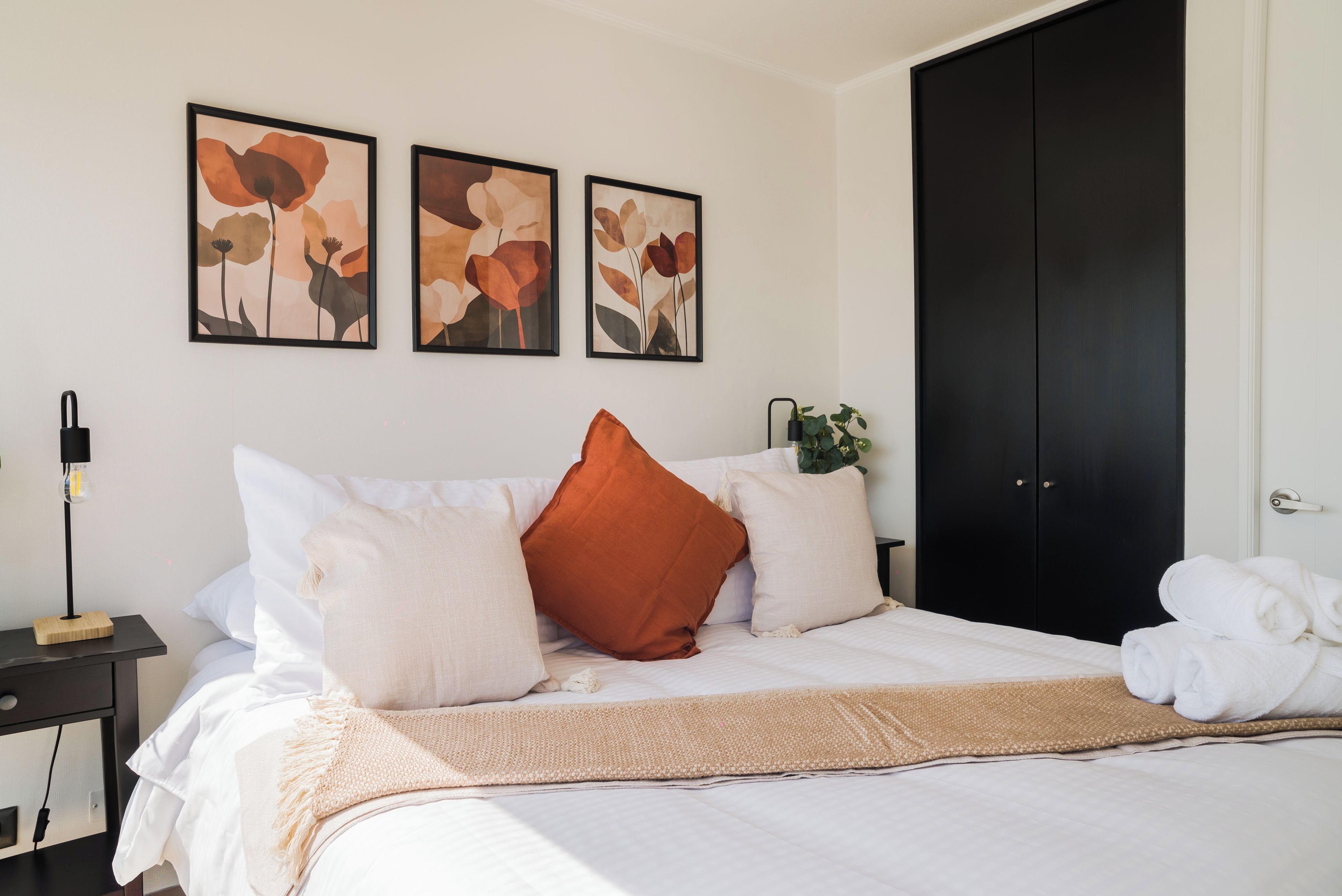 A neatly made bed with white bedding and a beige throw, a black wardrobe, and three framed floral artworks on the wall.