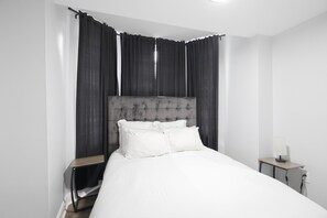A modern bedroom with a large bed, a nightstand, a lamp, and dark curtains. - Stylish 1bd Urban Haven Near Inner Harbor 1 Bedroom Apts (Baltimore)