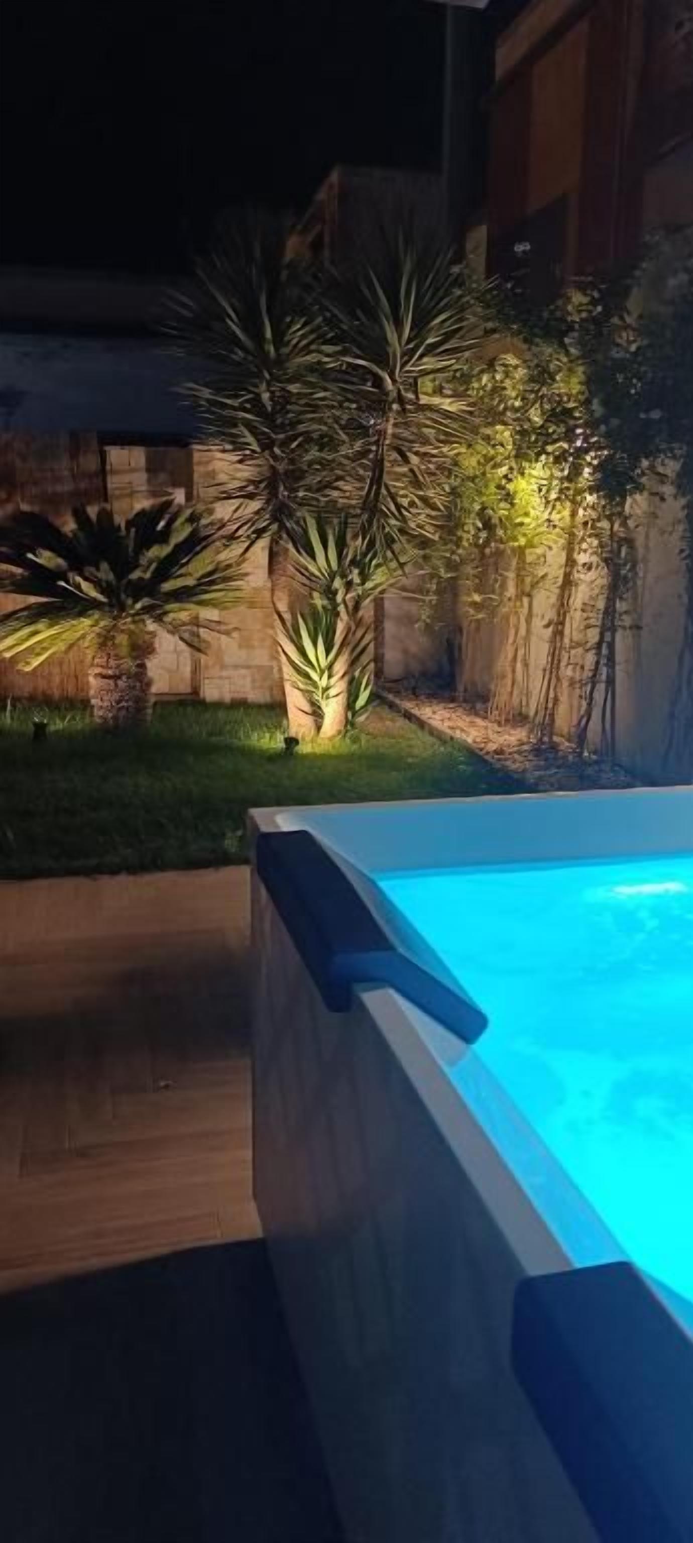 Una piscina con terraza de madera y un jardín iluminado por la noche.