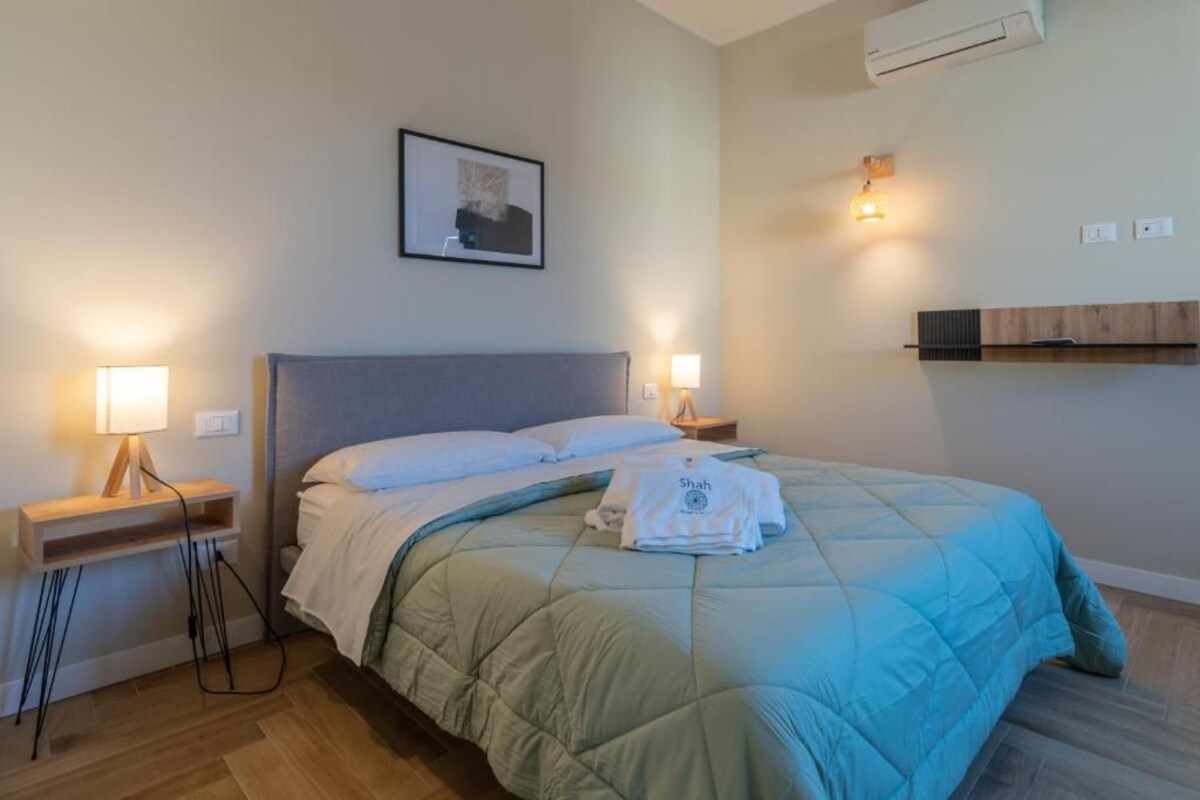 Hotellihuone, jossa on sänky, yöpöytävalaisimet, seinään asennettu ilmastointilaite ja sängyn yläpuolella oleva taulu.