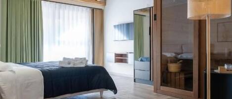 Habitación de hotel con piso de madera, una cama con cubrecama blanco, cortina verde y un ventanal grande.