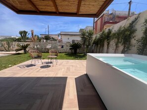 Un patio exterior con piscina, piso de madera y dos sillones de mimbre.