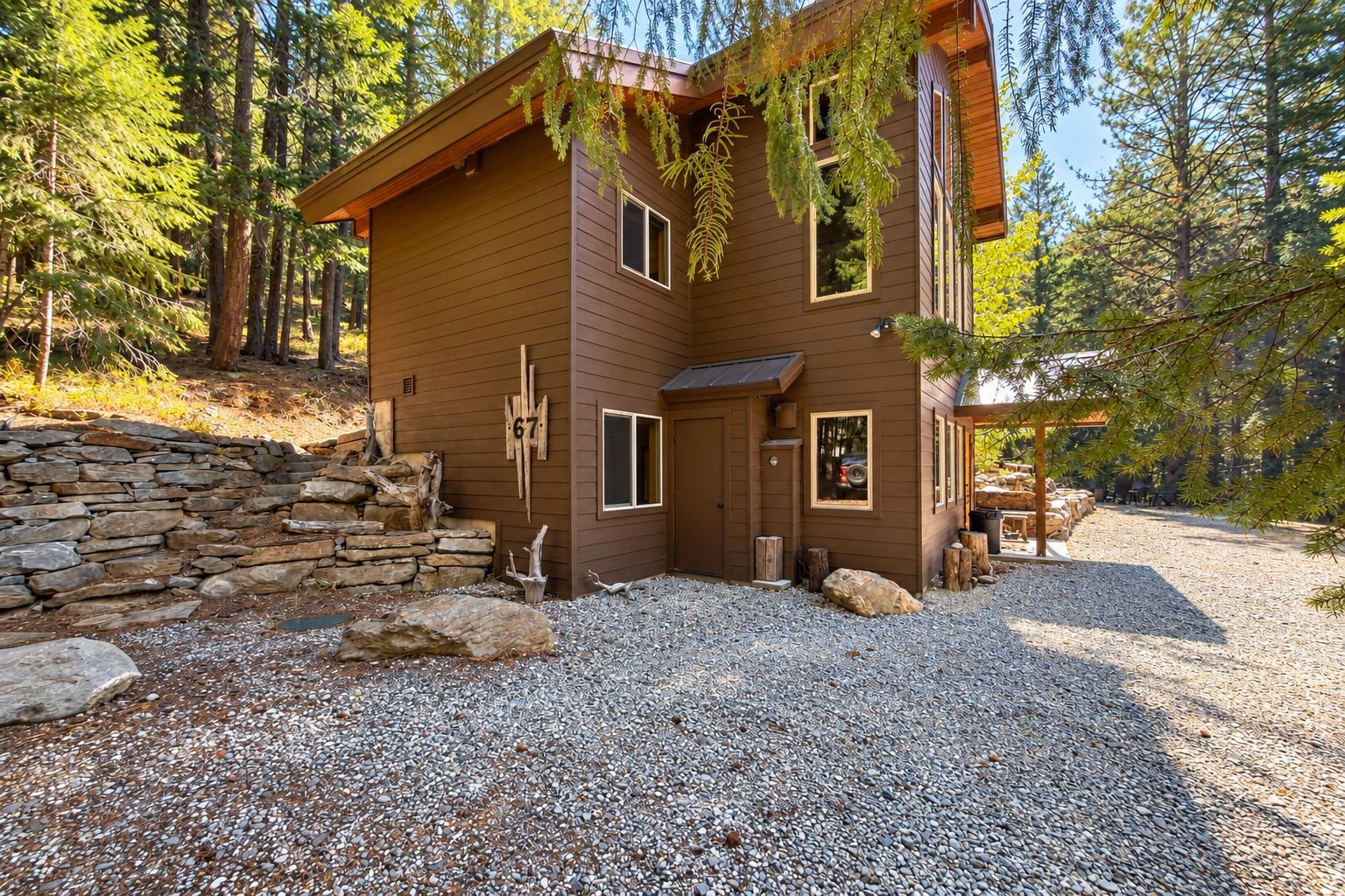 Une cabane dont l’entrée est encadrée par un mur de pierres, avec une allée en gravier et un décor de forêt en arrière-plan.