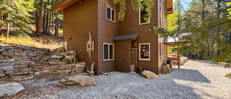 Une cabane dont l’entrée est encadrée par un mur de pierres, avec une allée en gravier et un décor de forêt en arrière-plan.