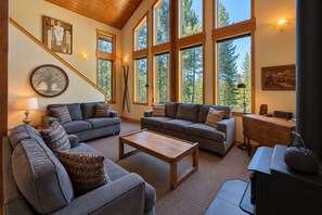 Ein gemütliches Wohnzimmer mit zwei Sofas, einem Couchtisch und einem Kamin. Große Fenster bieten Blick auf Bäume.