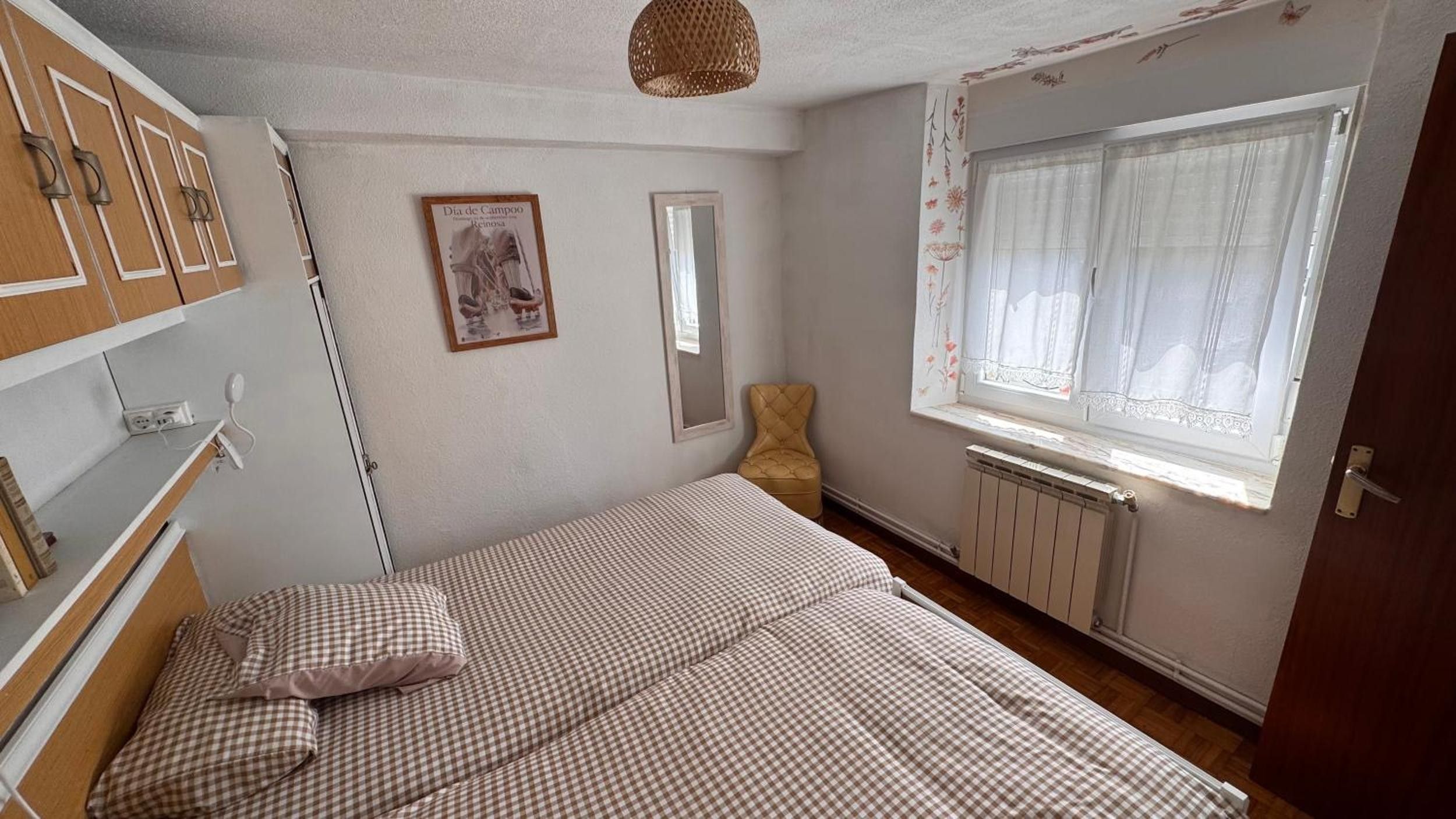 Un dormitorio con cama, una silla, un radiador, una ventana con cortinas y un estante montado en la pared.
