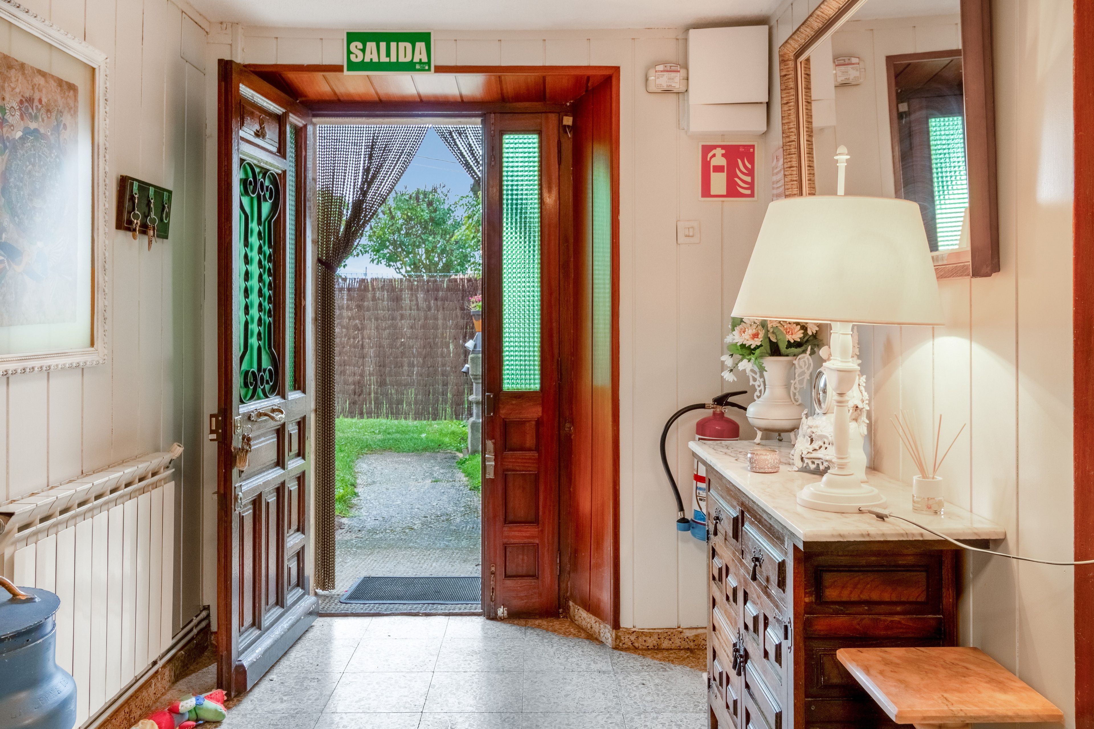 Passadís amb una porta de fusta que porta a l'exterior, amb la inscripció 'SALIDA', un radiador, una taula petita amb una làmpada i un extintor penjat a la paret.