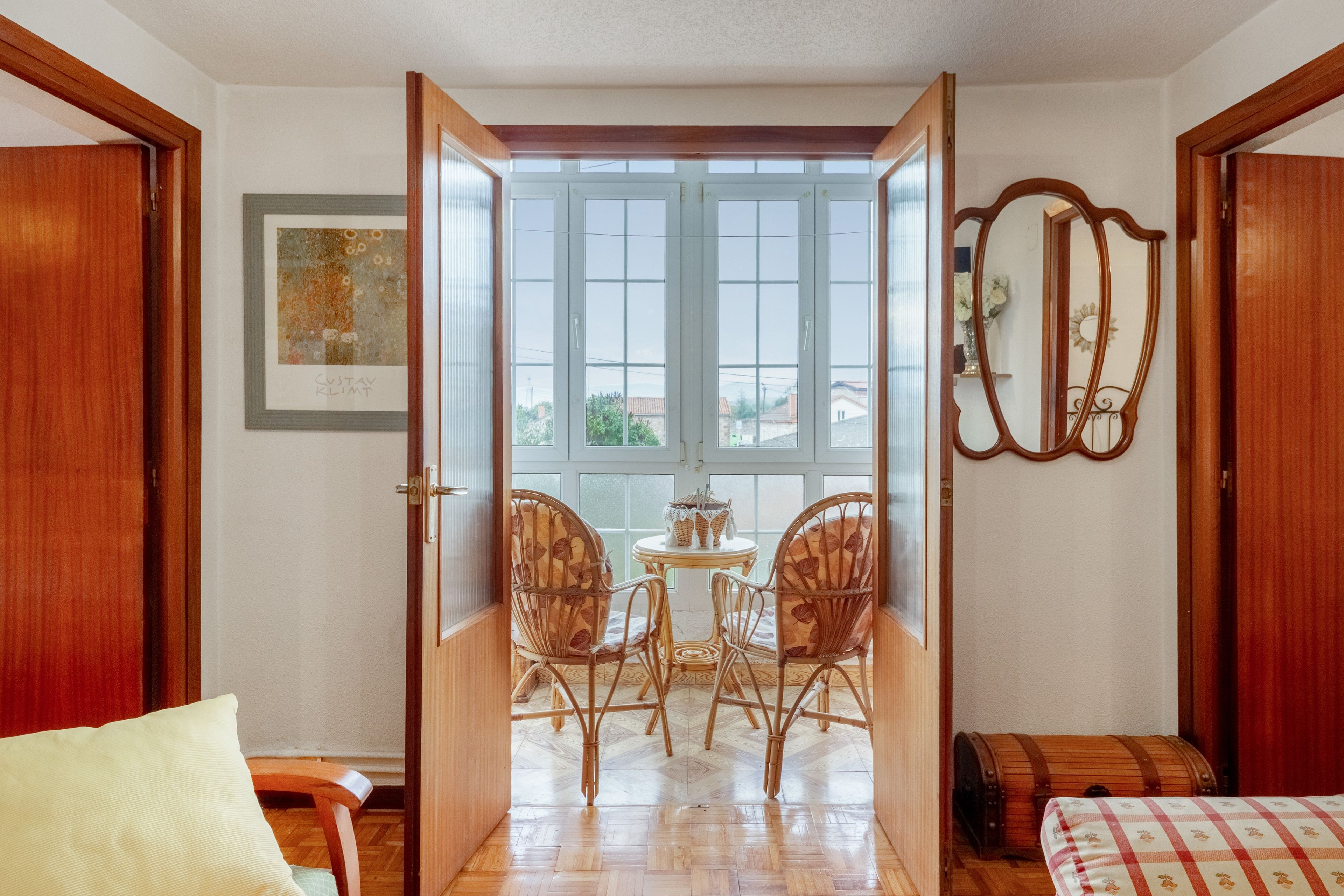 Una habitació amb una porta de fusta que dona a un balcó amb vistes, una taula petita amb cadires i un mirall a la paret.