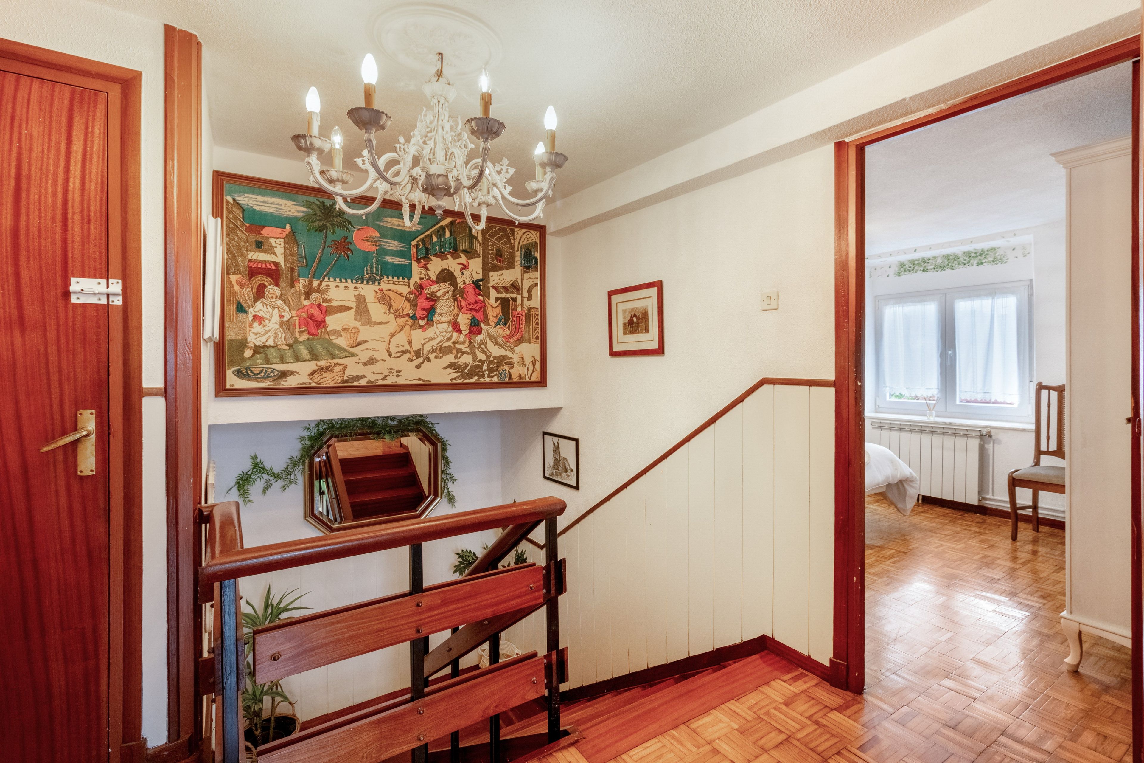 Una escalera con barandilla de madera, un candelabro y una pared con un tapiz grande y cuadros enmarcados.