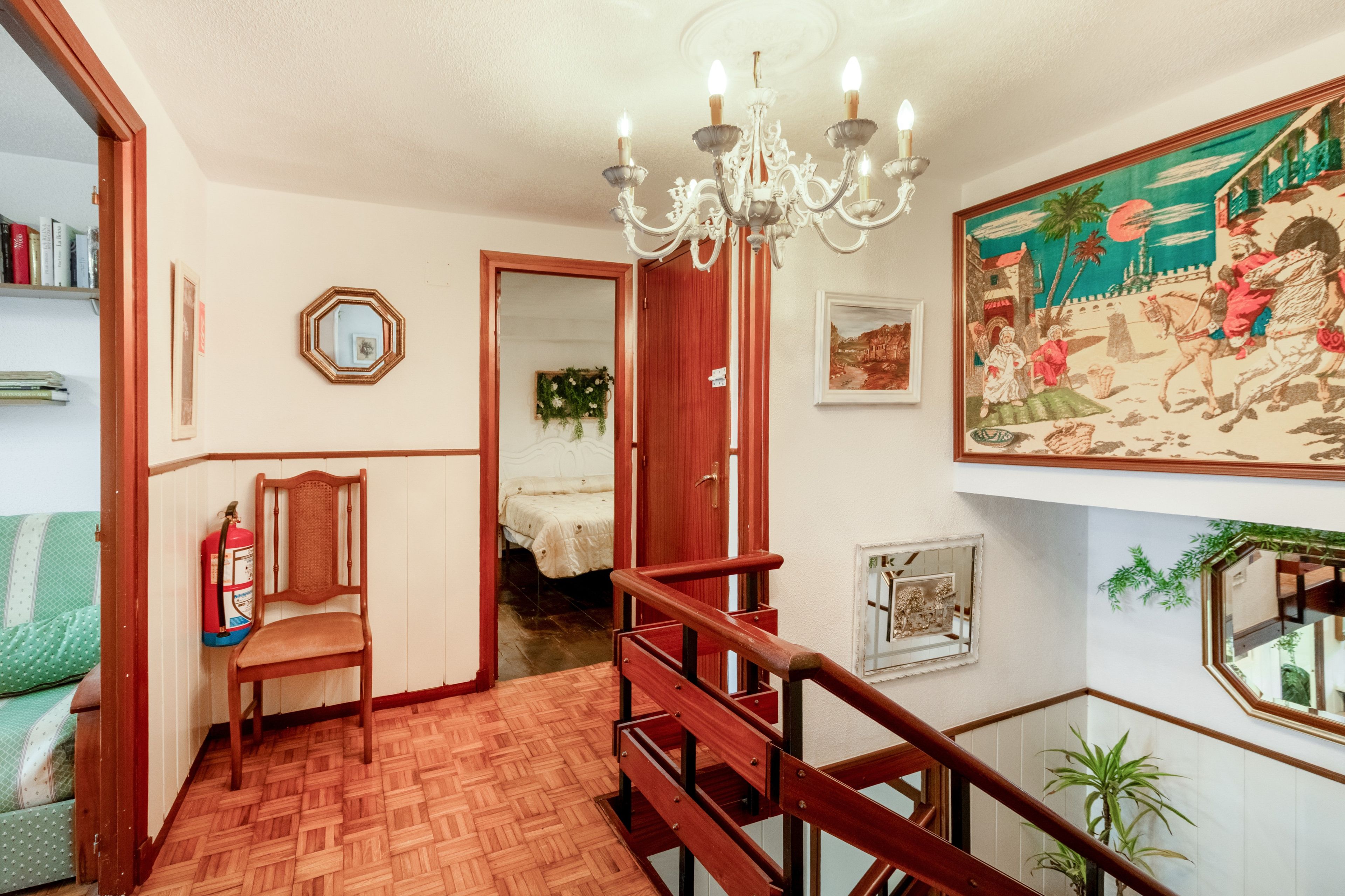 Une pièce avec un escalier en bois, un lustre, un lit, une chaise et un tableau accroché au mur.