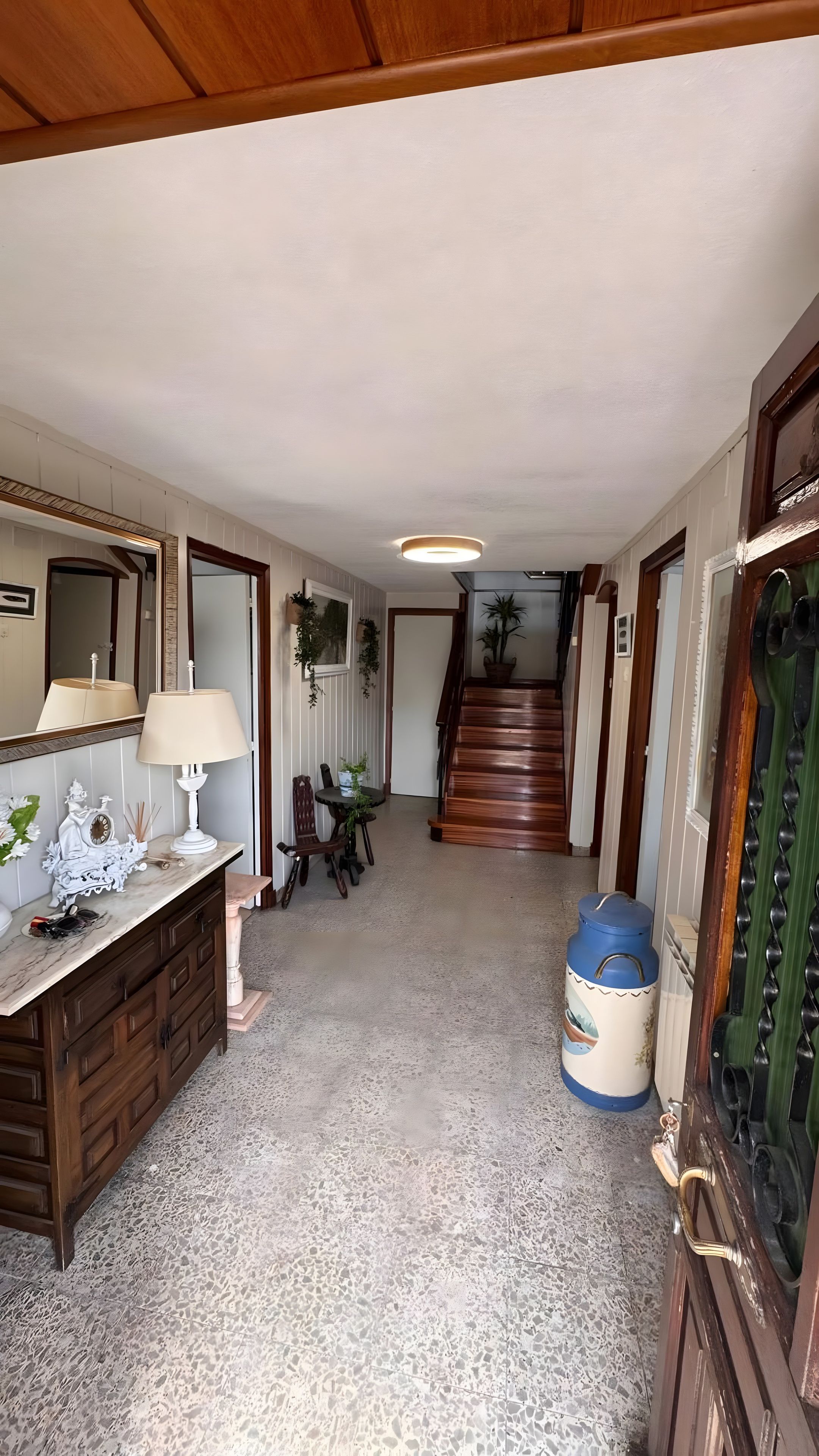 Un pasillo con una escalera de madera, un comedor con mesa y sillas, y un espejo grande a la izquierda.