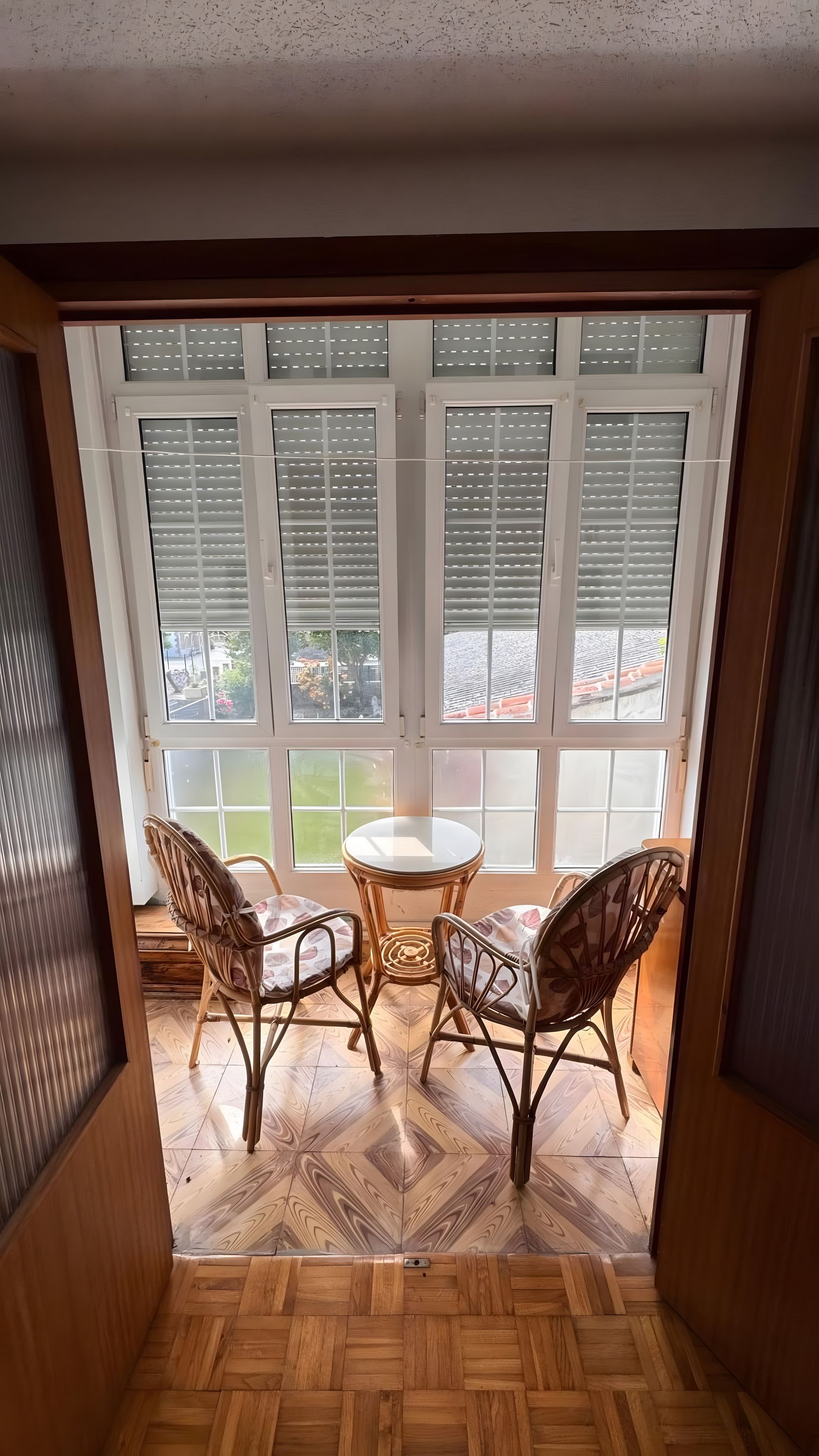 A room with a wooden herringbone floor, a round table with two chairs, and a view through a window with blinds.