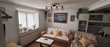 Ein gemütliches Wohnzimmer mit Sofa, Sessel und Couchtisch. Ein Fenster mit Vorhängen, ein Bild an der Wand und eine Kommode mit Dekorationselementen.