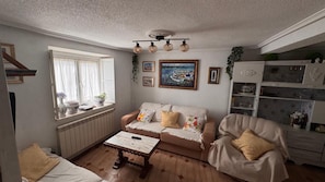 Ein gemütliches Wohnzimmer mit Sofa, Sessel und Couchtisch. Ein Fenster mit Vorhängen, ein Bild an der Wand und eine Kommode mit Dekorationselementen.