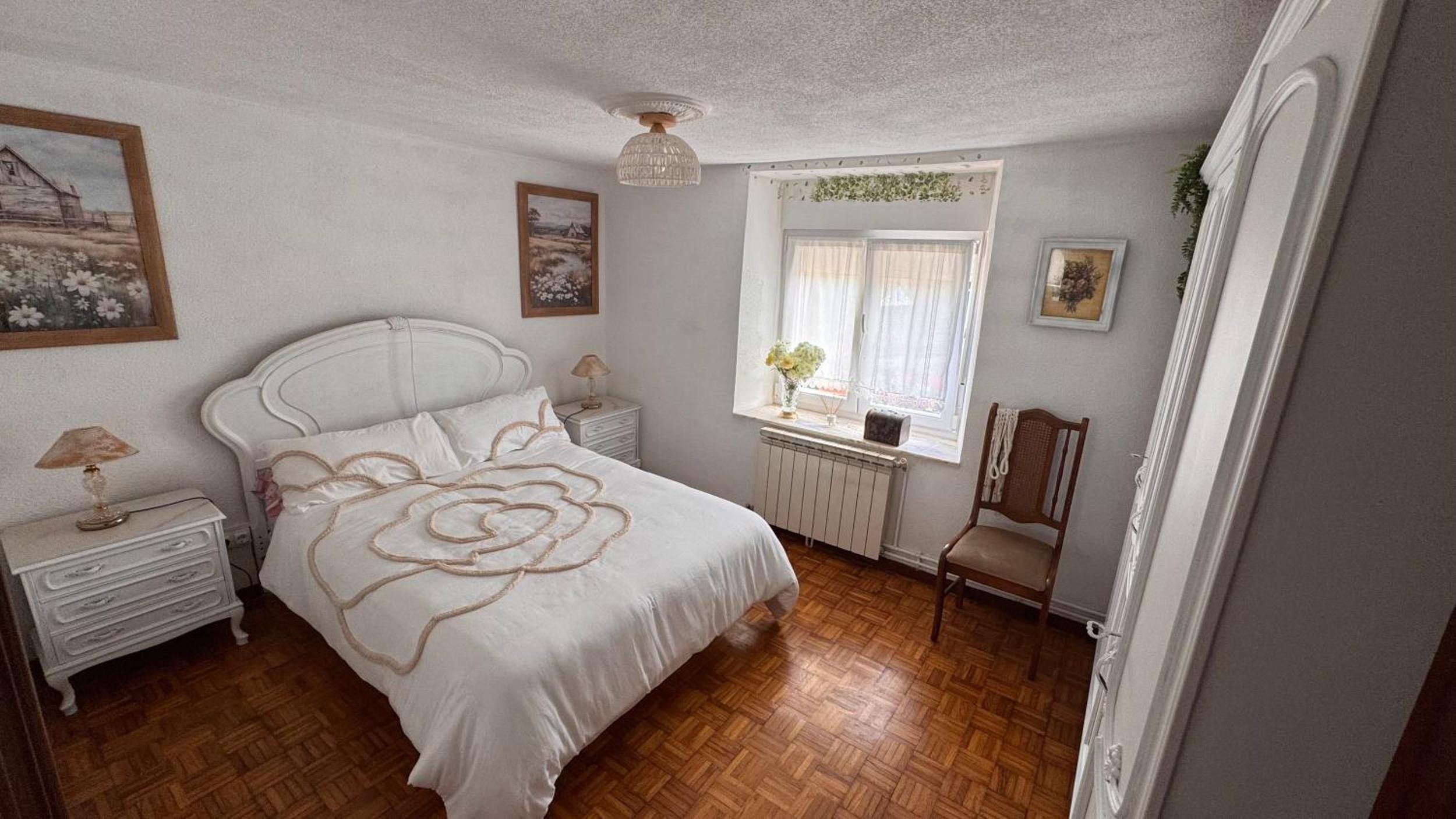 Une chambre à coucher avec un lit, des tables de chevet, une fenêtre avec des rideaux, un radiateur et une chaise.