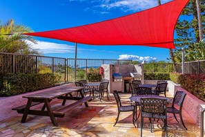 An outdoor patio with picnic tables and chairs, a barbecue grill, and a red shade sail. - Kihei Alii Kai C302 1 Bedroom Apts (Kihei)