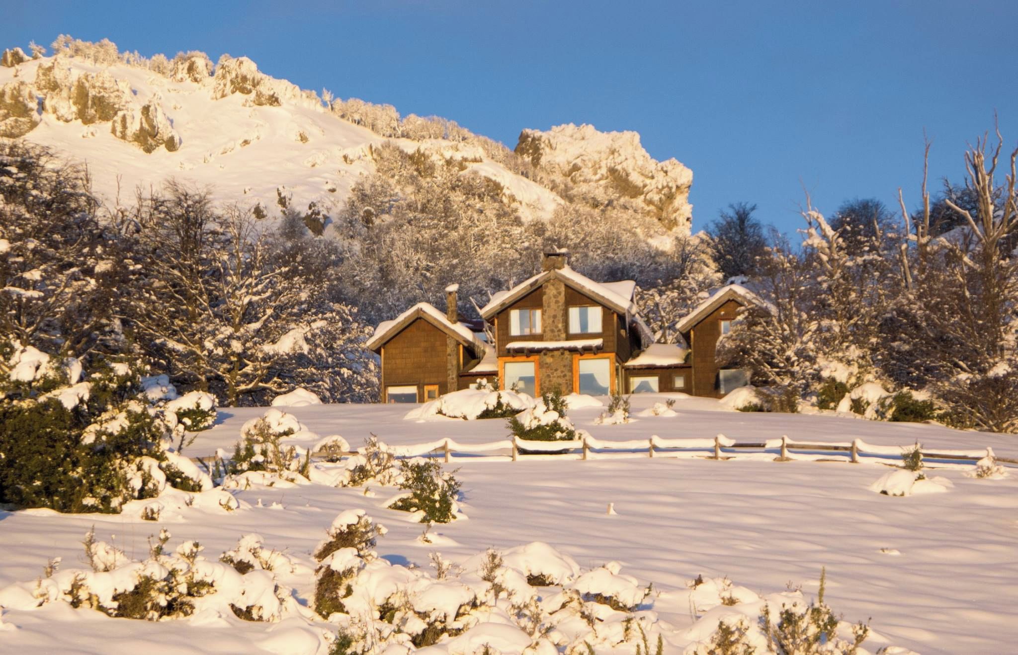 A snow-covered landscape with a wooden cabin and a fenced driveway.