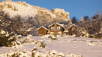 A snow-covered landscape with a wooden cabin and a fenced driveway.