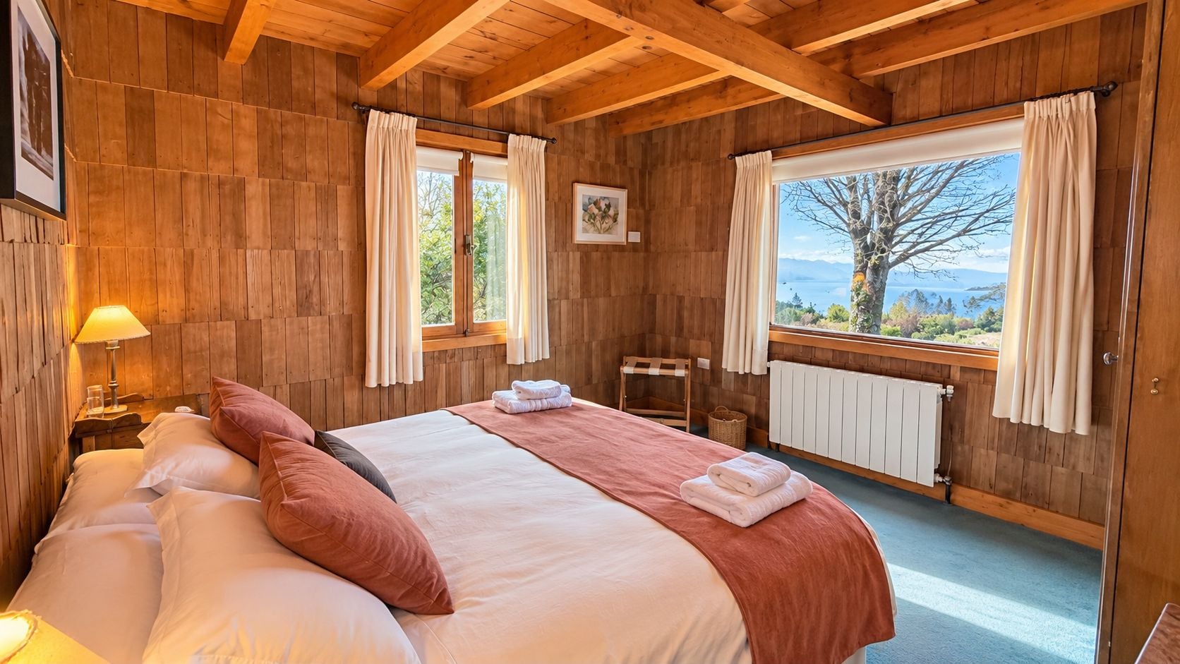 Un dormitorio de cabaña de madera con una cama, una mesita de noche, una lámpara, una ventana con cortinas y vistas al exterior.