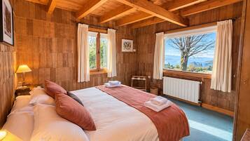 Un dormitorio de cabaña de madera con una cama, una mesita de noche, una lámpara, una ventana con cortinas y vistas al exterior.