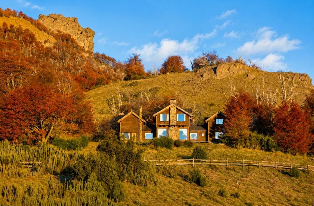 Une maison en bois à deux étages, dotée d’une cheminée en pierre, entourée d’arbres aux couleurs automnales et surplombant un ciel dégagé.