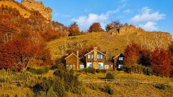 Une maison en bois à deux étages, dotée d’une cheminée en pierre, entourée d’arbres aux couleurs automnales et surplombant un ciel dégagé.