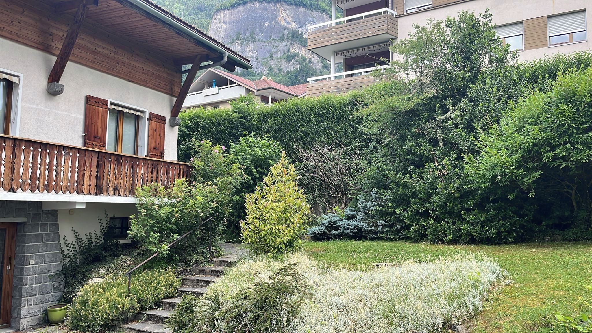A residential building with a wooden balcony, stone steps, and a lush garden with various plants and flowers.