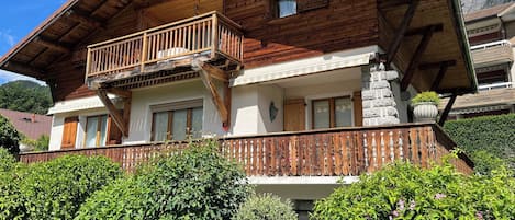 A two-story wooden chalet with a balcony, surrounded by lush greenery.